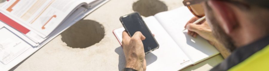 Urban roofer taking notes holding iPhone and writing in an urban location wearing hard hat and safety jacket