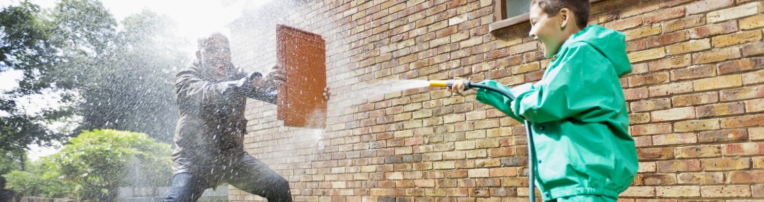 Boy hosing man holding a clay roof tile for protection in front of brick-built façade
